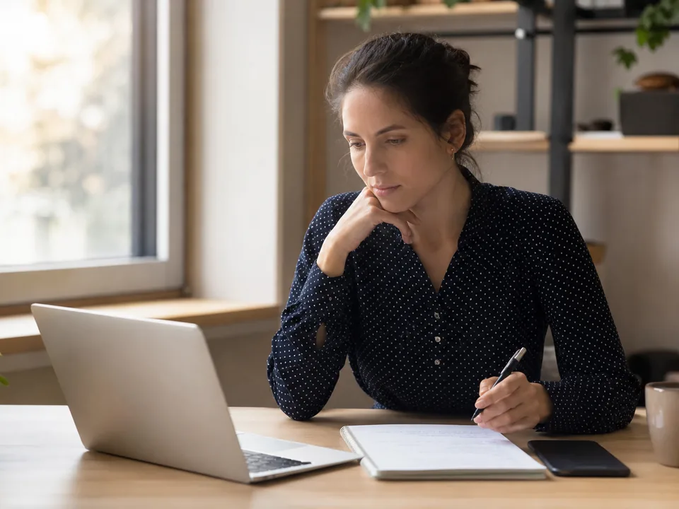 A woman at a desk in the morning comparing two app subscriptions on a laptop and a phone with a notepad of sums