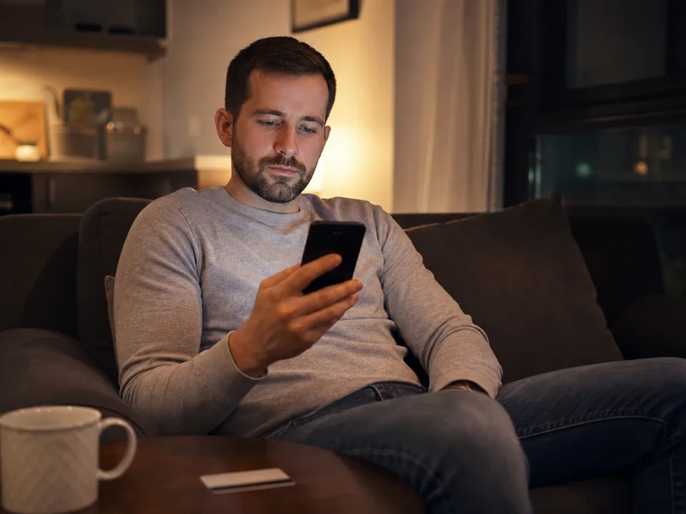 A man on his sofa in the evening checking a subscription price on his phone with a card beside him