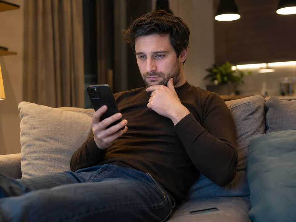 A man on his couch in the evening looking at a subscription checkout on his phone with a card beside him