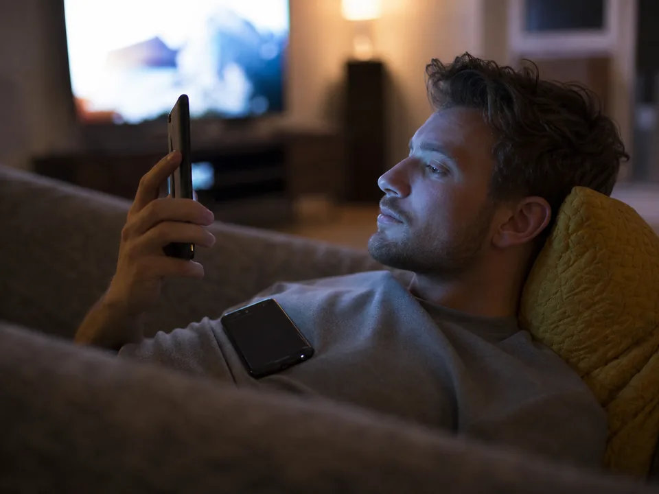 A man on a couch at night reading a long chat on his phone with a second phone resting beside him